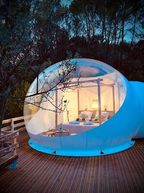 A transparent dome structure with a bed and a table inside, surrounded by a wooden deck and trees. - La Villa Tosca (Callas)