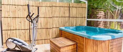 A hot tub with a wooden bench, a stationary bike, and a bamboo fence in a covered outdoor area.