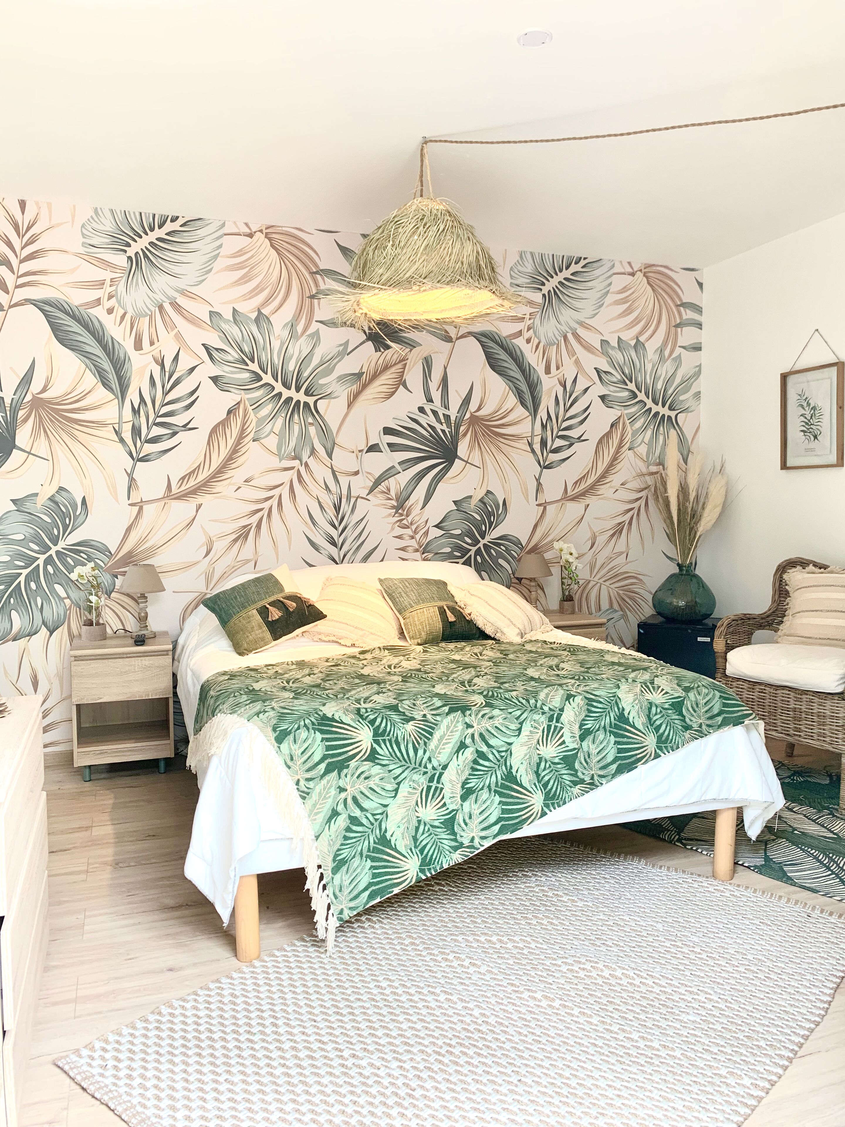 Een slaapkamer met een bed, een nachtkastje, een rieten stoel, een hanglamp en een muur met een tropisch bladpatroon.