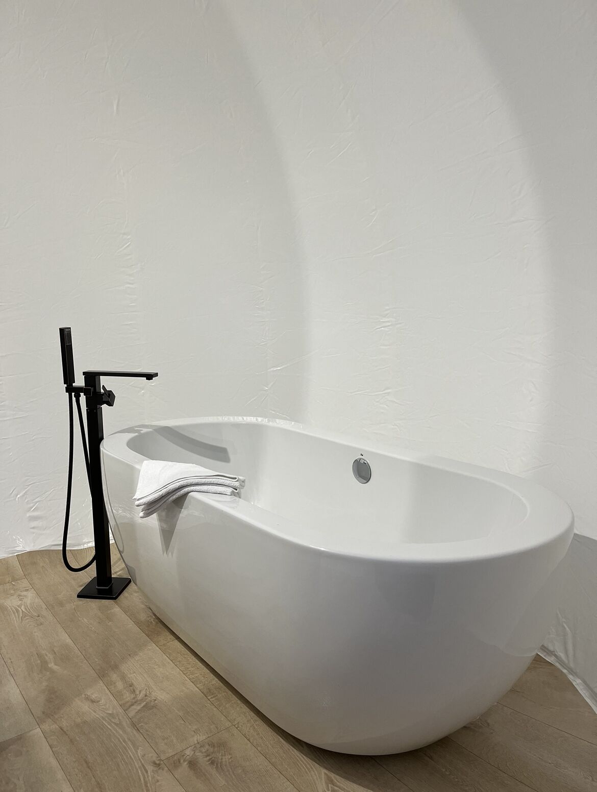 A modern, freestanding bathtub in a minimalist bathroom with a wooden floor and plain white walls.