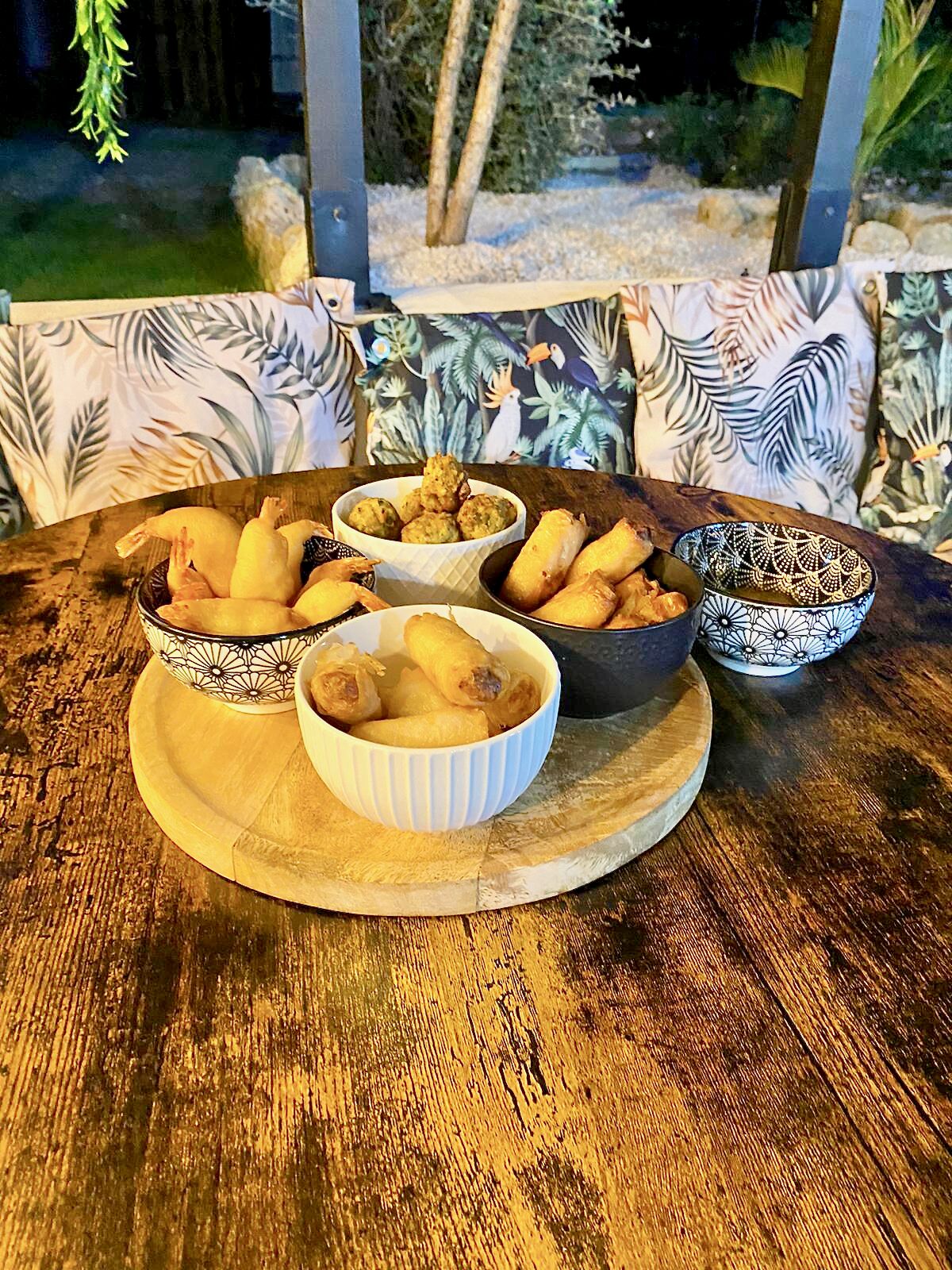 A wooden table with bowls of food, likely a meal, set on a patio with patterned cushions and a view of trees and a fence.