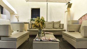 Uma área de estar moderna ao ar livre, com móveis de vime, uma mesa central com revistas e um grande guarda-sol para proteção do sol.