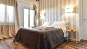 Uma cama bem arrumada com um edredom escuro, uma mesinha de cabeceira com abajur e uma janela com cortinas.