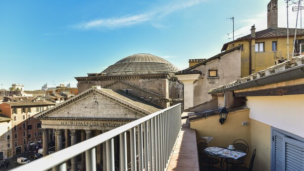 Uma varanda com mesa e cadeiras, com vista para um edifício histórico com colunas e cúpula.