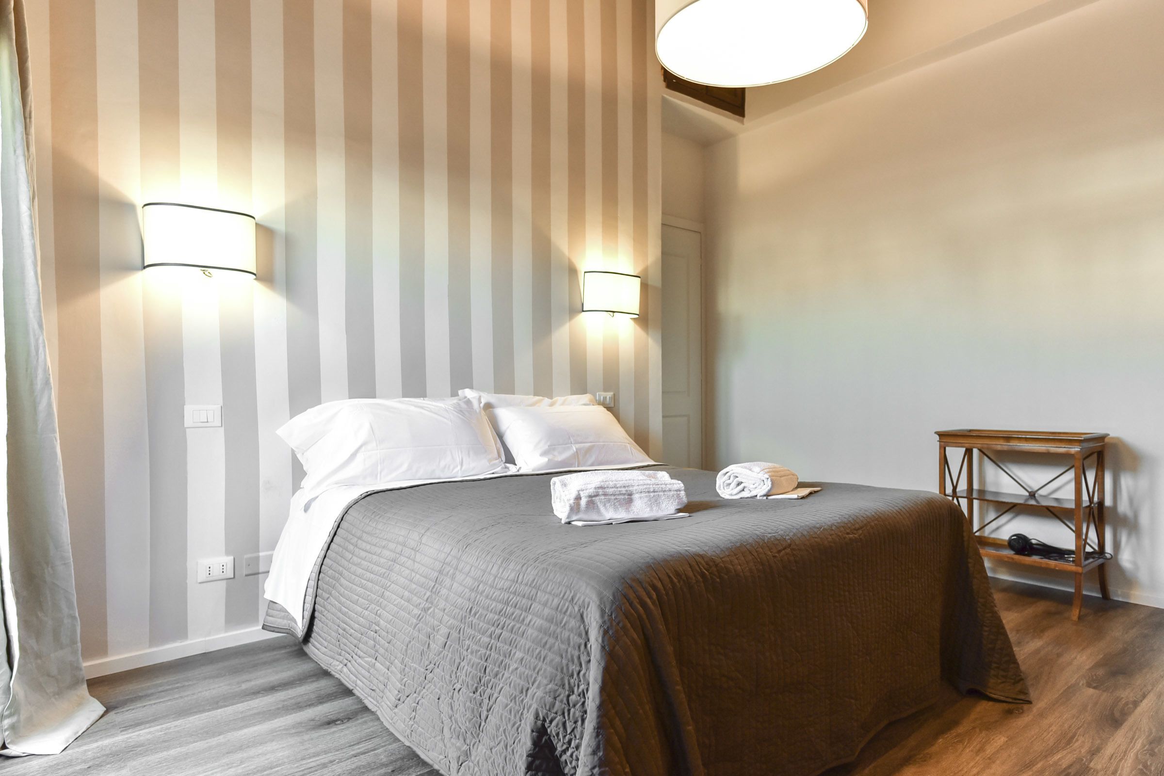 A neatly made bed with a grey quilt, two bedside lamps, and a wooden nightstand with a lamp.