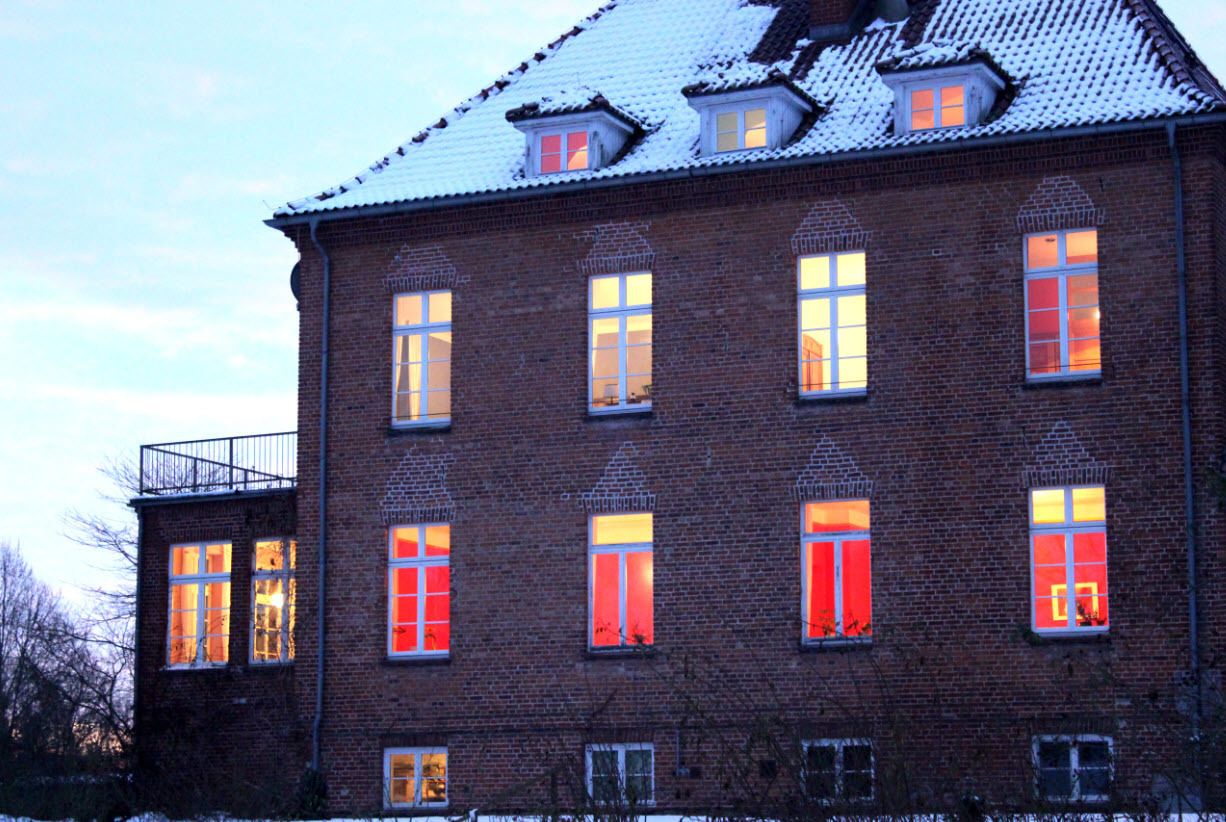 Ein Backsteinhaus mit mehreren Fenstern, von denen einige rote Fensterläden haben, und einem Balkon an der linken Seite.