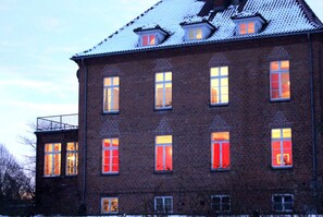 Un bâtiment en briques doté de nombreuses fenêtres, dont certaines sont agrémentées de volets rouges, et d’un balcon situé sur le côté gauche.