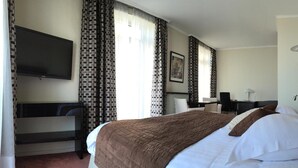 A hotel room with a bed, a desk, a chair, a TV, and patterned curtains. - Savoy Hôtel Evian (Evian-les-Bains)