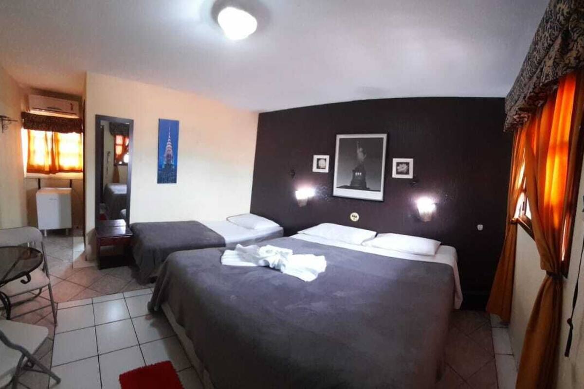 A hotel room with two beds, a small table, and a chair. There is a window with curtains, a wall-mounted air conditioner, and a door with a curtain.