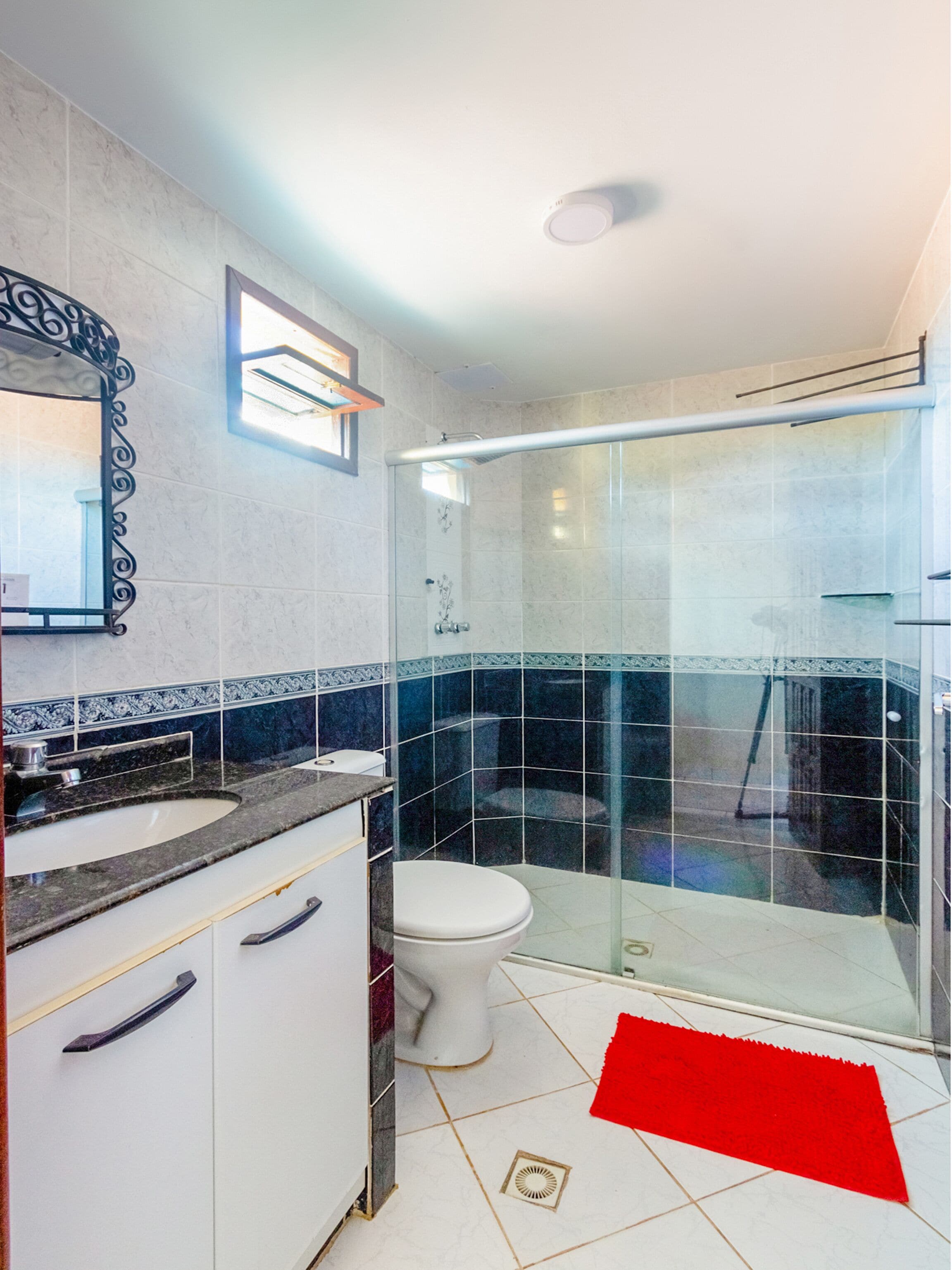 Une salle de bain avec une douche à parois de verre, des toilettes blanches, un lavabo avec un plan de toilette en marbre et un tapis rouge sur le sol.