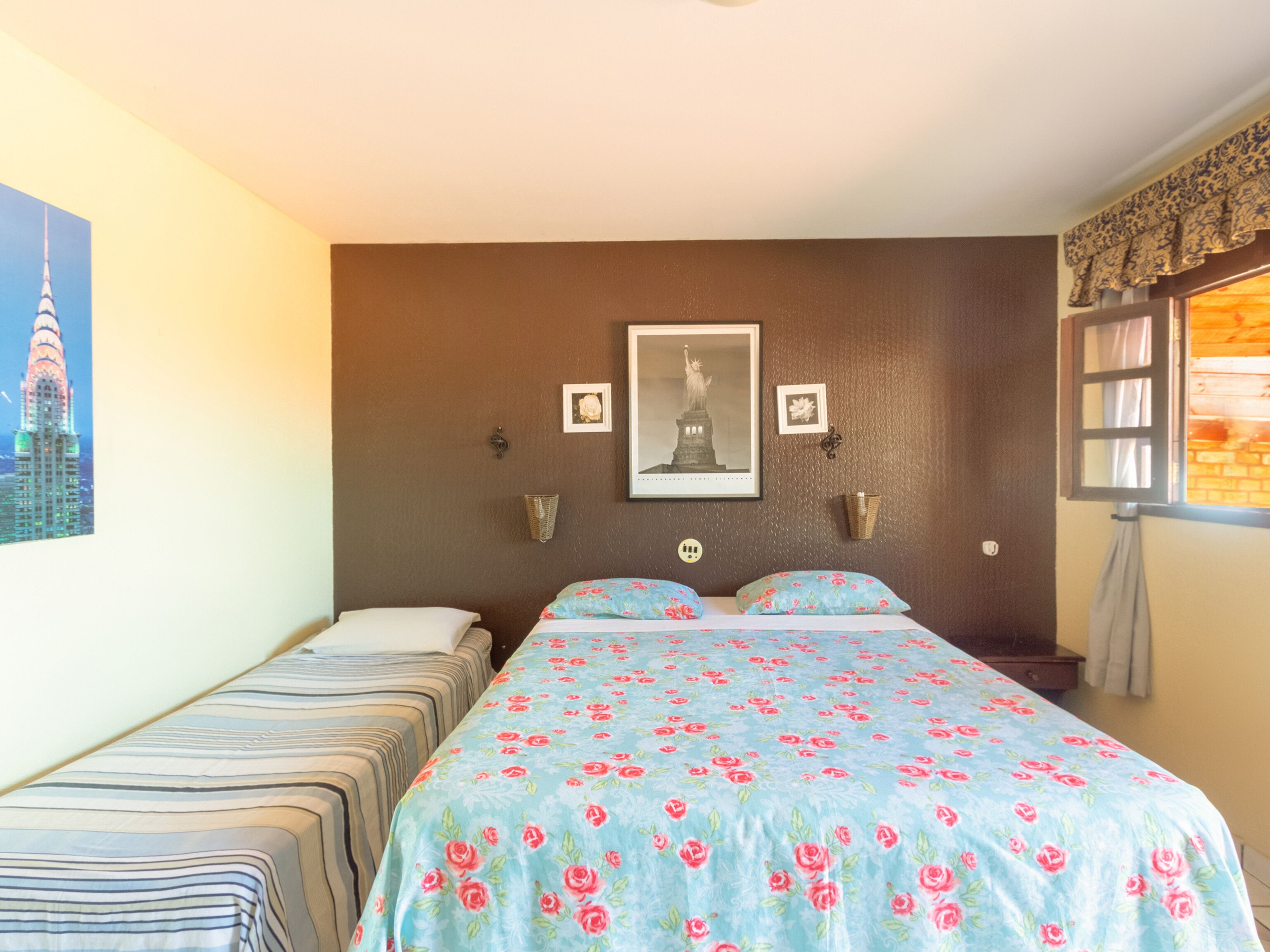 Habitación de hotel con dos camas individuales, una cama grande con cubrecama floral, un cuadro con la Estatua de la Libertad y una ventana con cortinas.