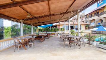 An outdoor patio with wooden tables and chairs, a thatched roof, and a view of a pool and buildings in the background.