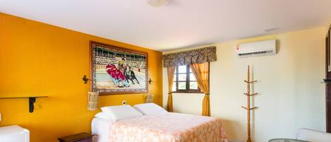 A bedroom with a bed, a bedside table, a chair, a small table, a wall-mounted air conditioner, and a window with curtains.
