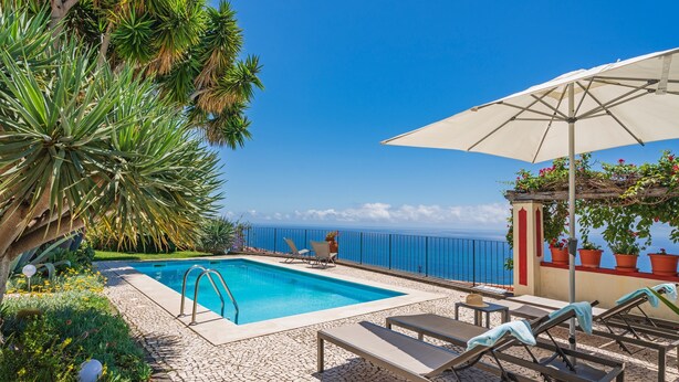 A pool area with lounge chairs and an umbrella, surrounded by palm trees and a view of the sea. - Quinta D'Alegria by Our Madeira (Funchal)