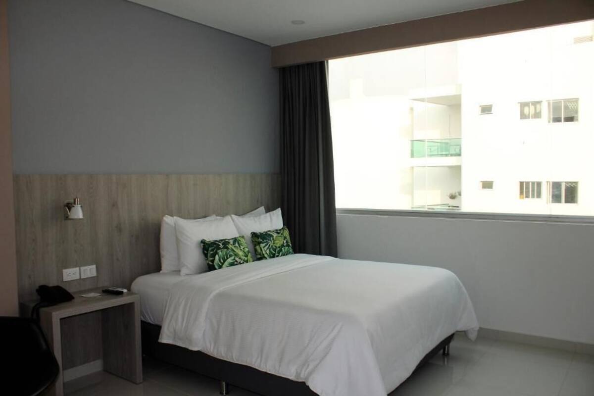 Una habitación de hotel con una cama grande, cabecera de madera, mesita de noche, escritorio y ventana con vista a un edificio.