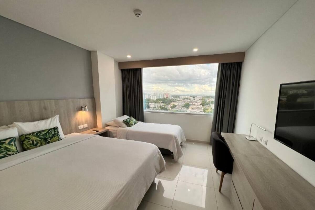 Habitación de hotel con dos camas, un escritorio, un televisor y un amplio ventanal con vistas a la ciudad.