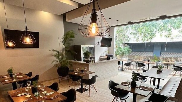 Restaurante moderno com mesas de madeira e cadeiras pretas, uma televisão grande e vista para o verde lá fora.