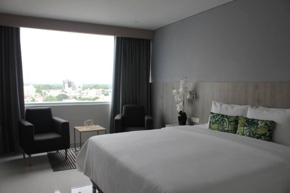 Una habitación de hotel moderna con una cama grande, dos sillones, una mesita y vistas a la ciudad a través de amplios ventanales.