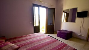 Un dormitorio con una cama, ventana, puerta, televisión y un pequeño taburete morado.