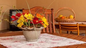 Una mesa con mantel floral, una planta en maceta con flores rojas y amarillas, y una cesta de fruta sobre la mesa.