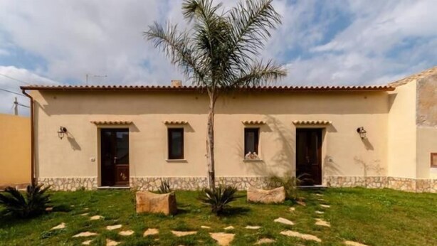 Un edificio de dos pisos con techo plano, una entrada central y un pequeño jardín con una palmera y rocas decorativas.