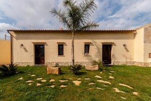 A two-story building with a flat roof, a central entrance, and a small garden with a palm tree and decorative rocks.