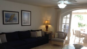 A living room with a dark blue sofa, a wicker chair, a glass coffee table, and two framed pictures on the wall. - Holiday Home at 163 Port St Charles (Speightstown)