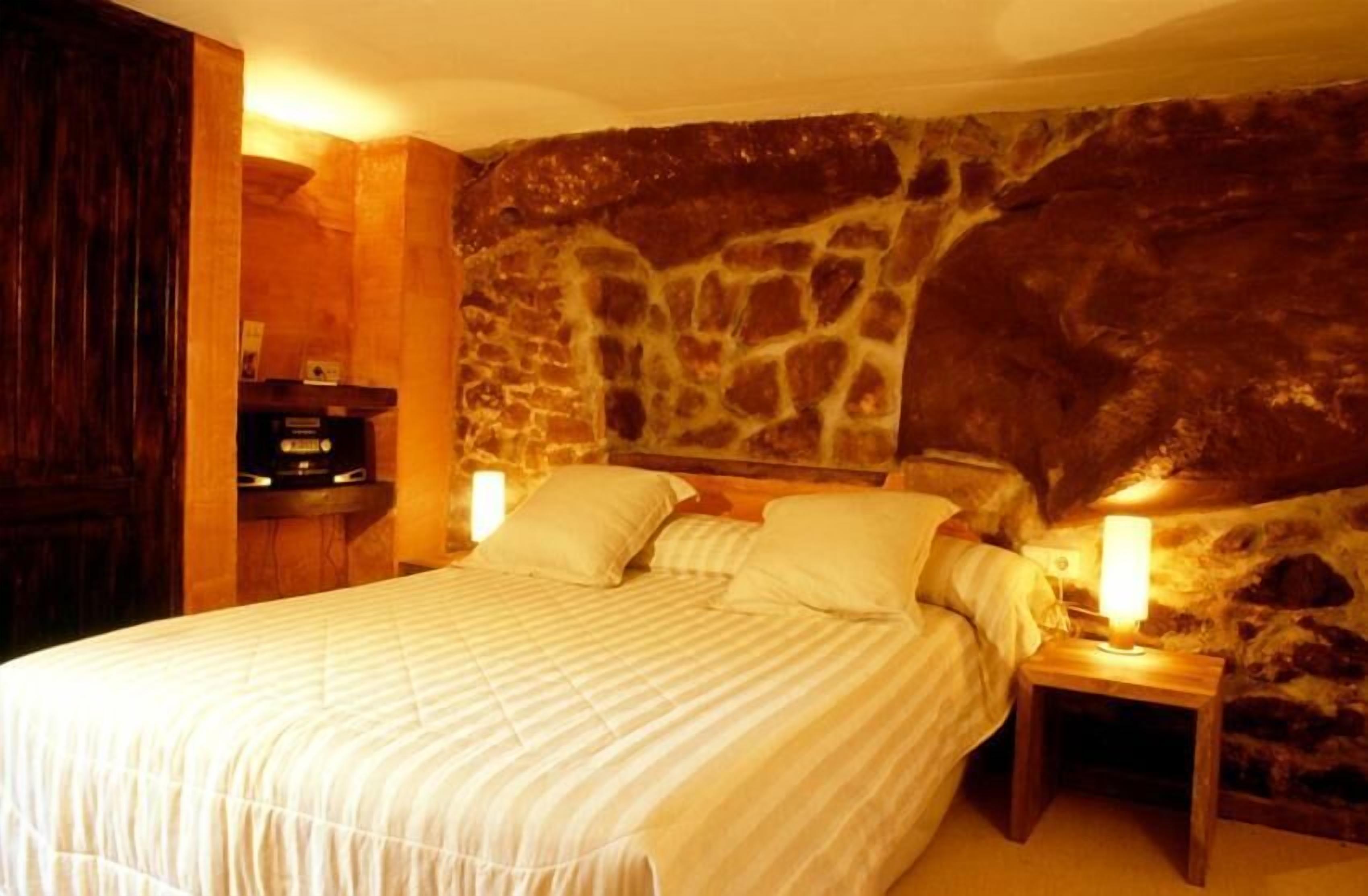 A bedroom with a stone wall, a bed with two pillows, and a nightstand with a lamp.