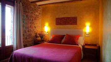 A bedroom with a stone wall, a bed with red bedding, two bedside lamps, and a nightstand with a vase of flowers.