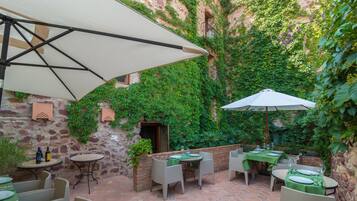 An outdoor patio area with brick flooring, green tables, and chairs, surrounded by lush greenery and stone walls.
