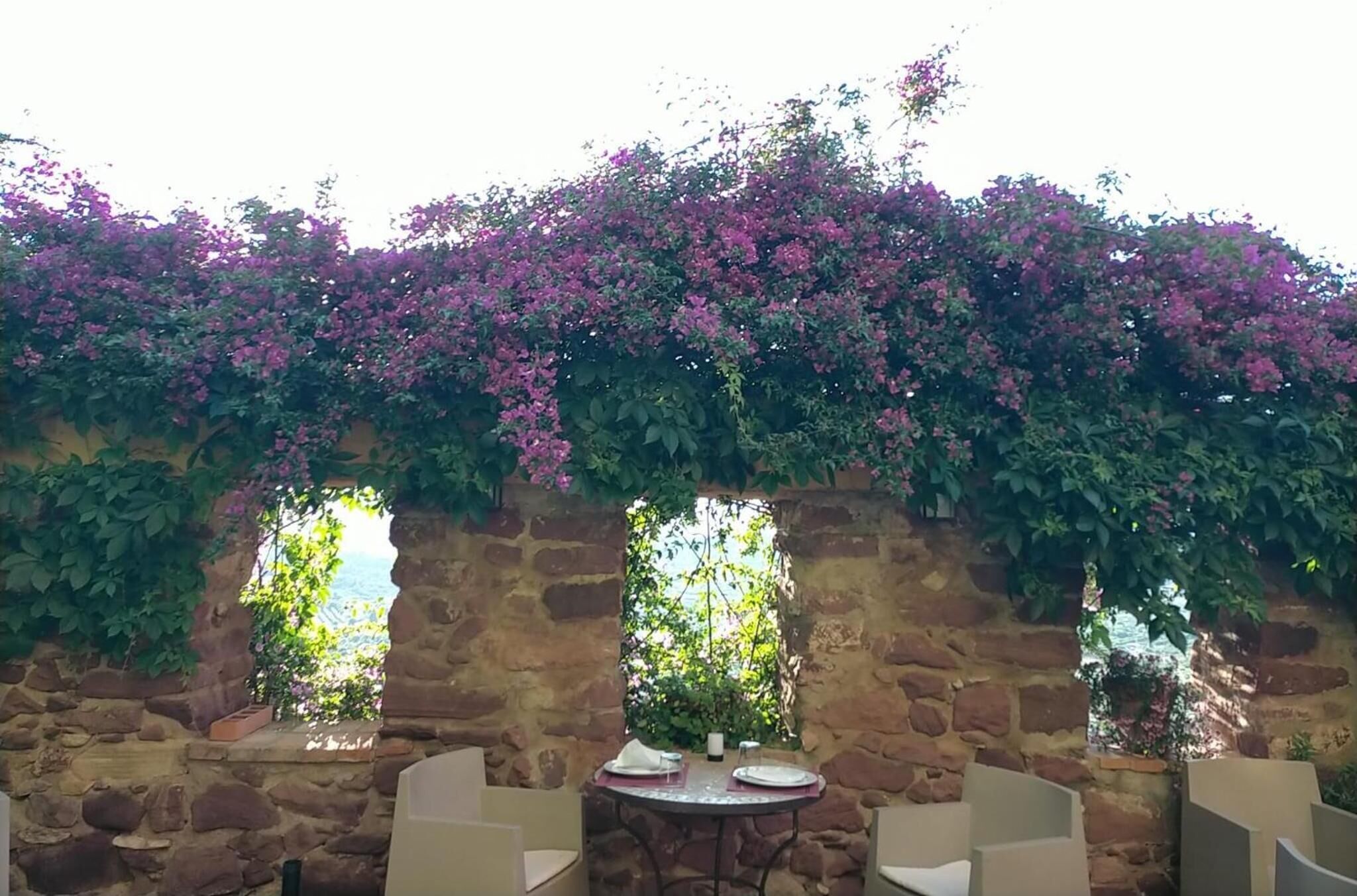 A stone wall with a table and chairs, covered by flowering vines.