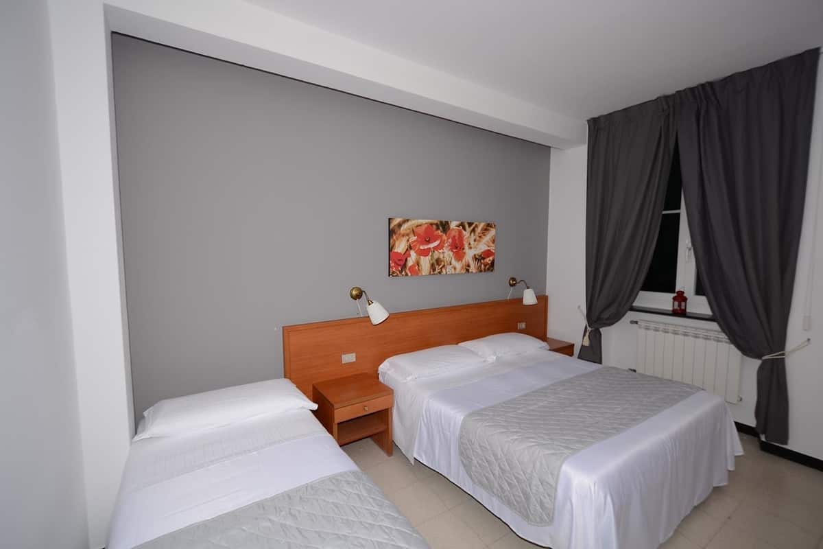 A hotel room with two single beds, a wooden headboard, a nightstand, a wall-mounted lamp, a window with curtains, and a radiator.