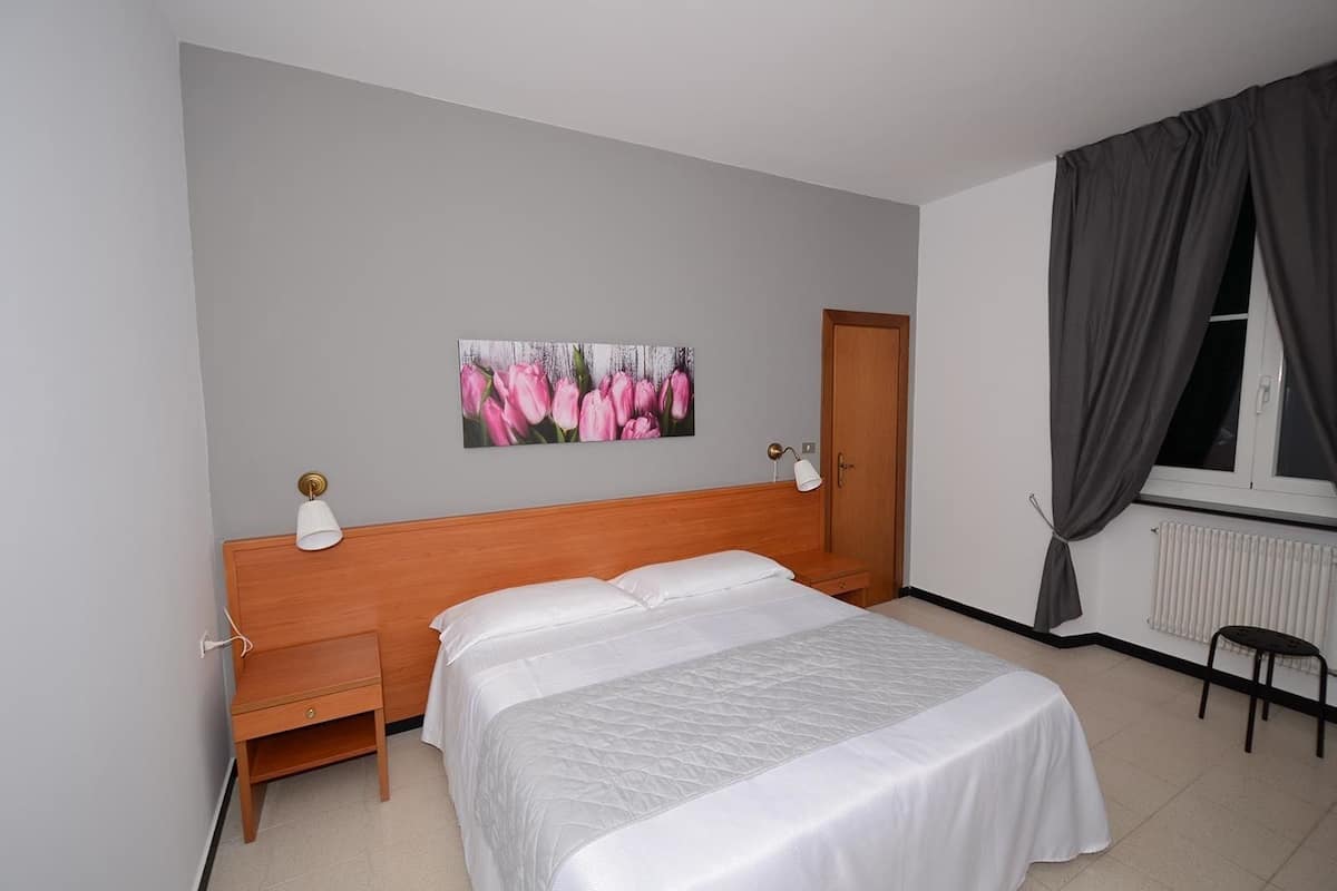 A bedroom with a bed, nightstands, a wooden headboard, a wall-mounted lamp, a radiator, and a window with curtains.