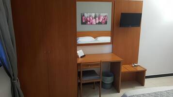 Ein Schlafzimmer mit einem Holzschrank, einem Schreibtisch mit Stuhl, einem Bett, einer kleinen Mülltonne und einem an der Wand befestigten Fernseher.