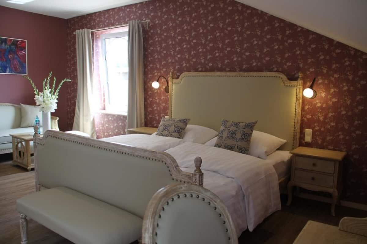 A bedroom with a large bed, two bedside tables, a nightstand, a bench, and a window with curtains.