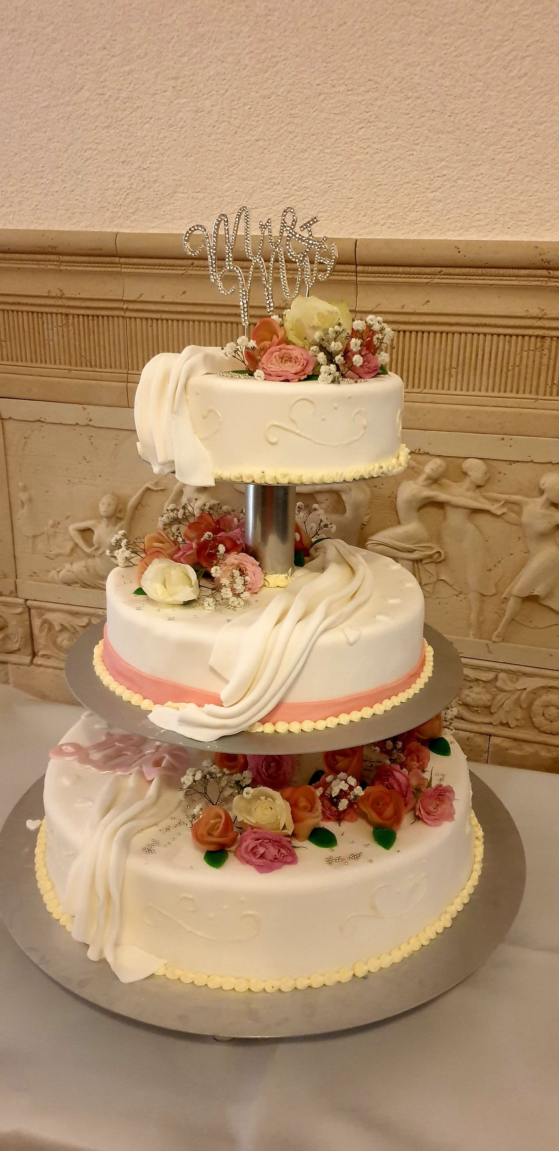 Um bolo de casamento em vários andares, decorado com flores e um enfeite prateado.