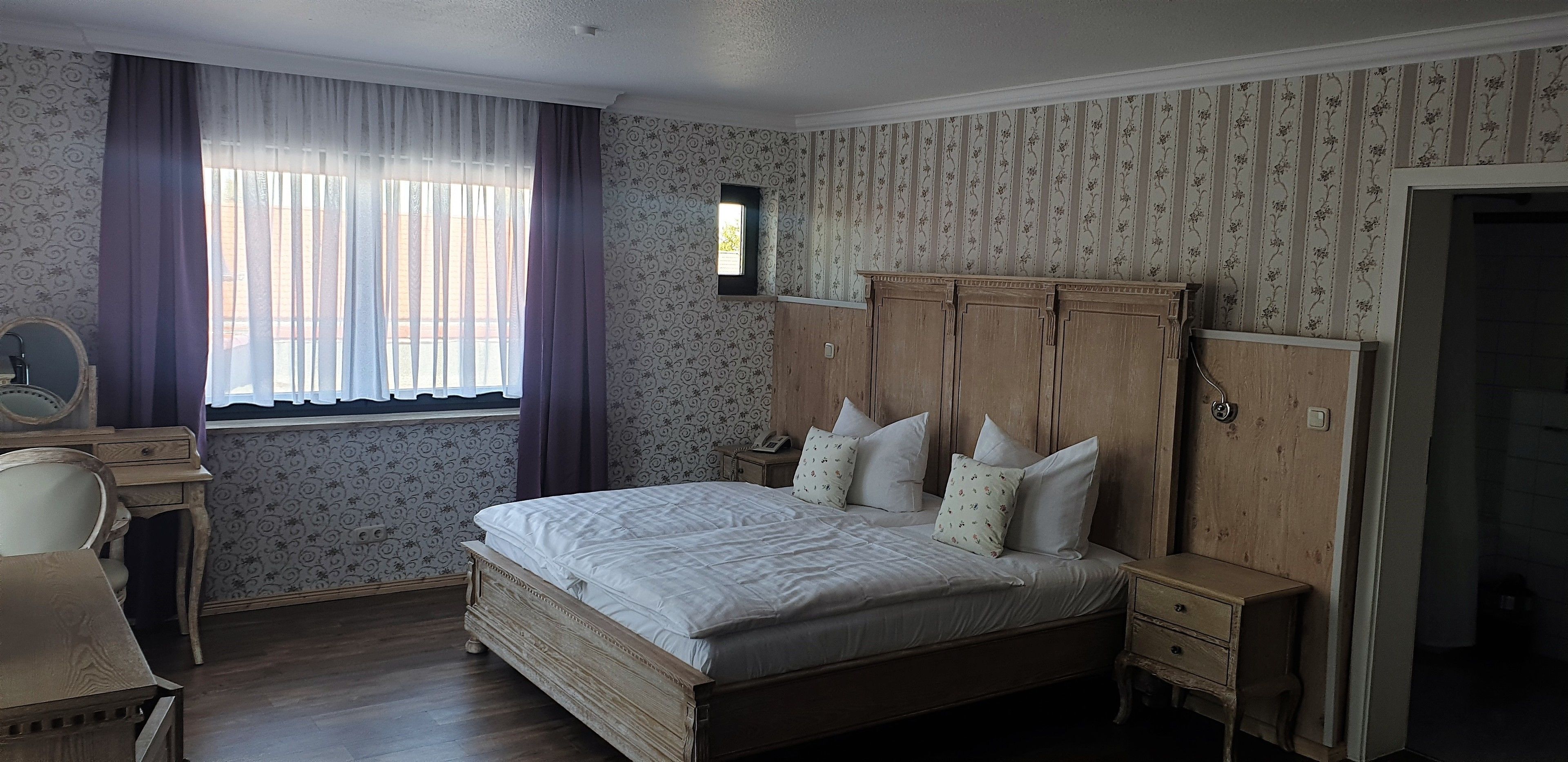A bedroom with a wooden bed, a desk, a chair, a wardrobe, and a window with curtains.