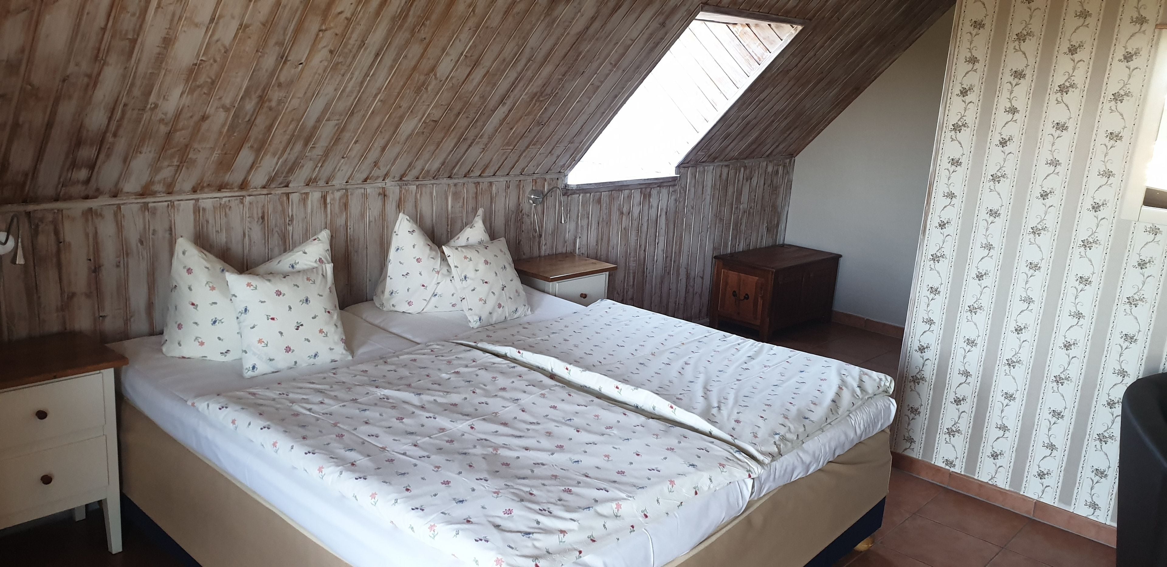 A bedroom with a sloped ceiling, a skylight, a bed with floral bedding, a nightstand, a wooden cabinet, and a patterned curtain.