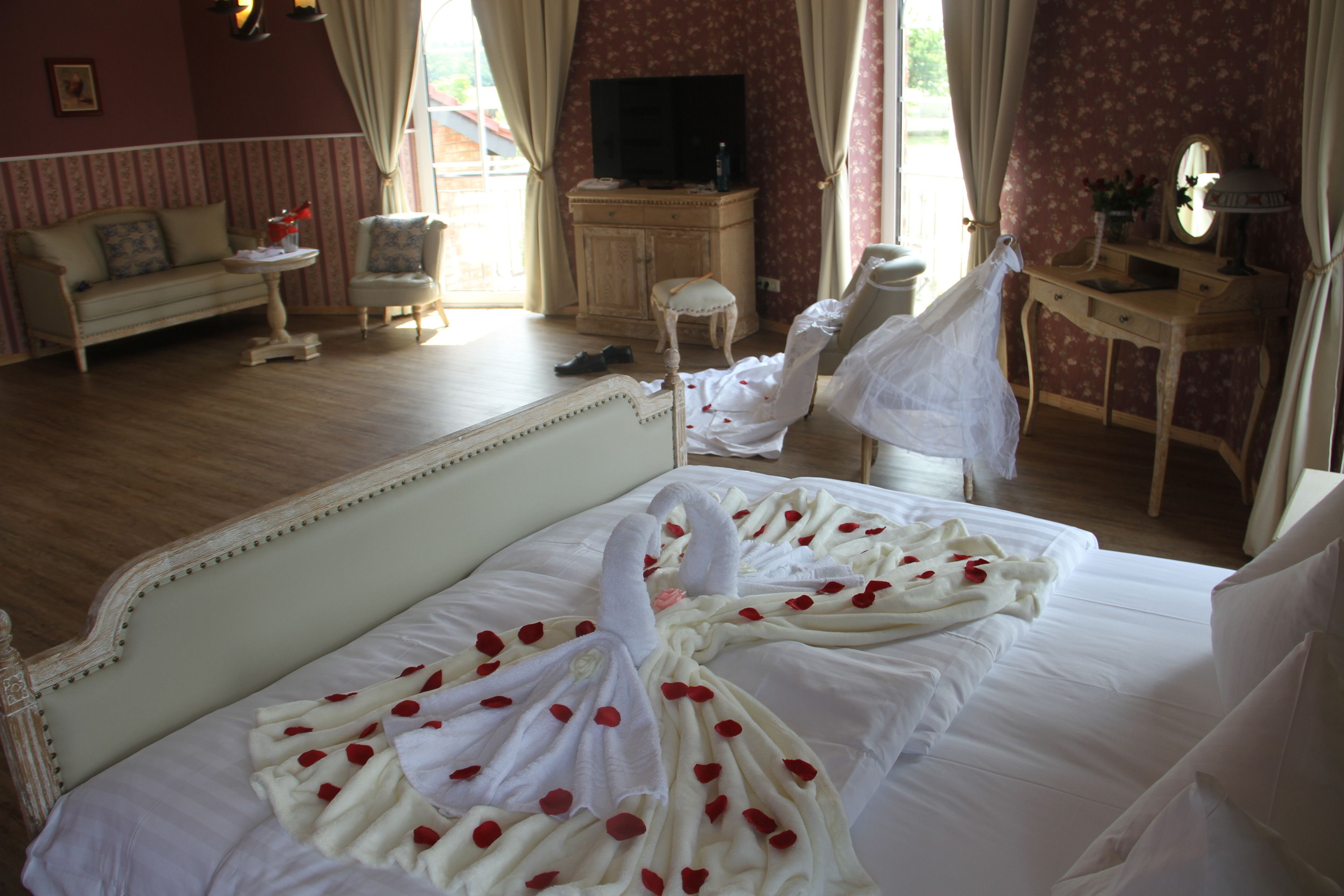 Habitación de hotel con una cama decorada con pétalos de rosa, un televisor, un tocador con espejo y una pared cubierta de papel tapiz floral.