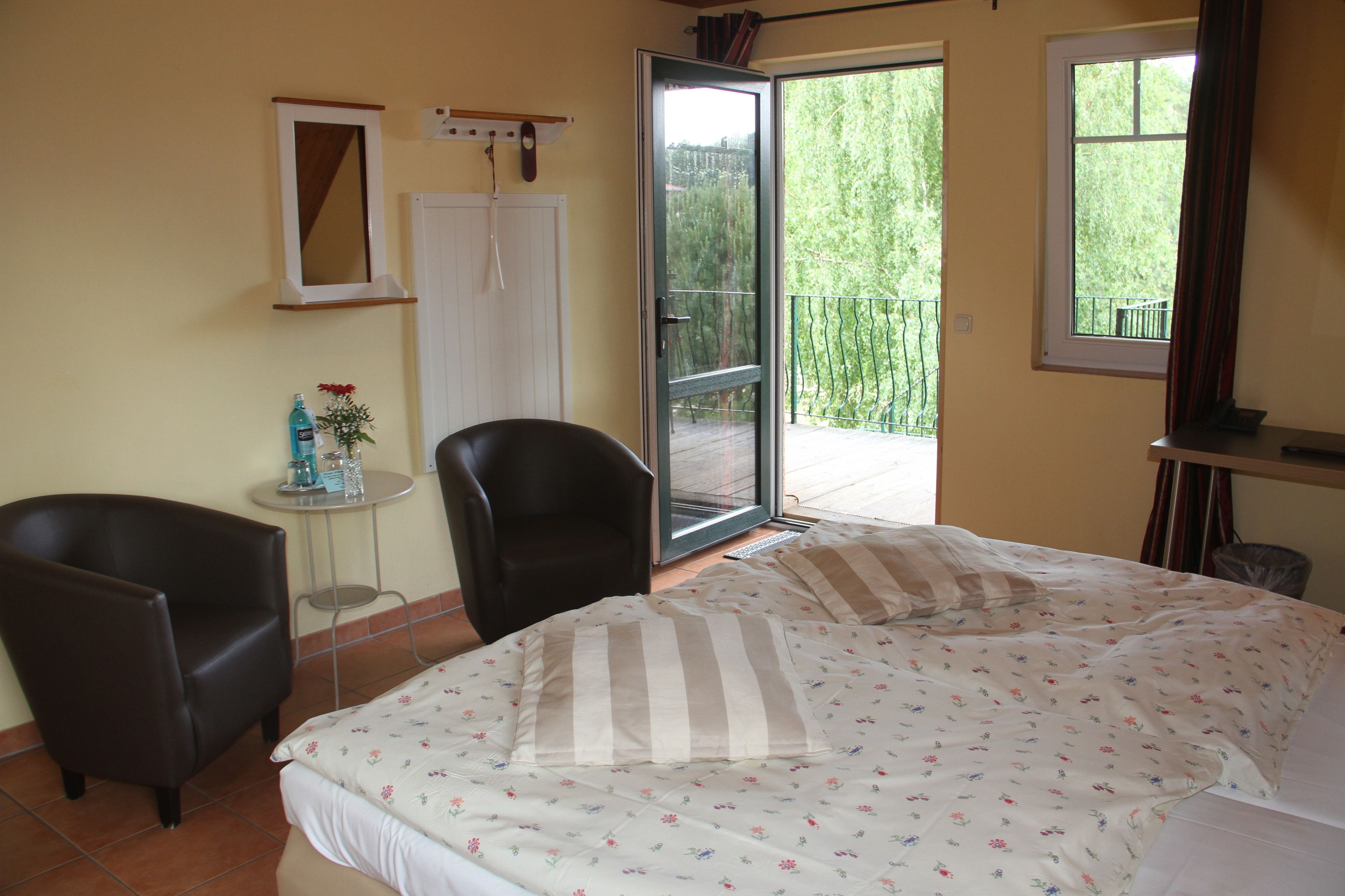 Une chambre à coucher avec un lit, deux chaises, une petite table sur laquelle se trouvent une bouteille et des fleurs, une porte et une fenêtre.