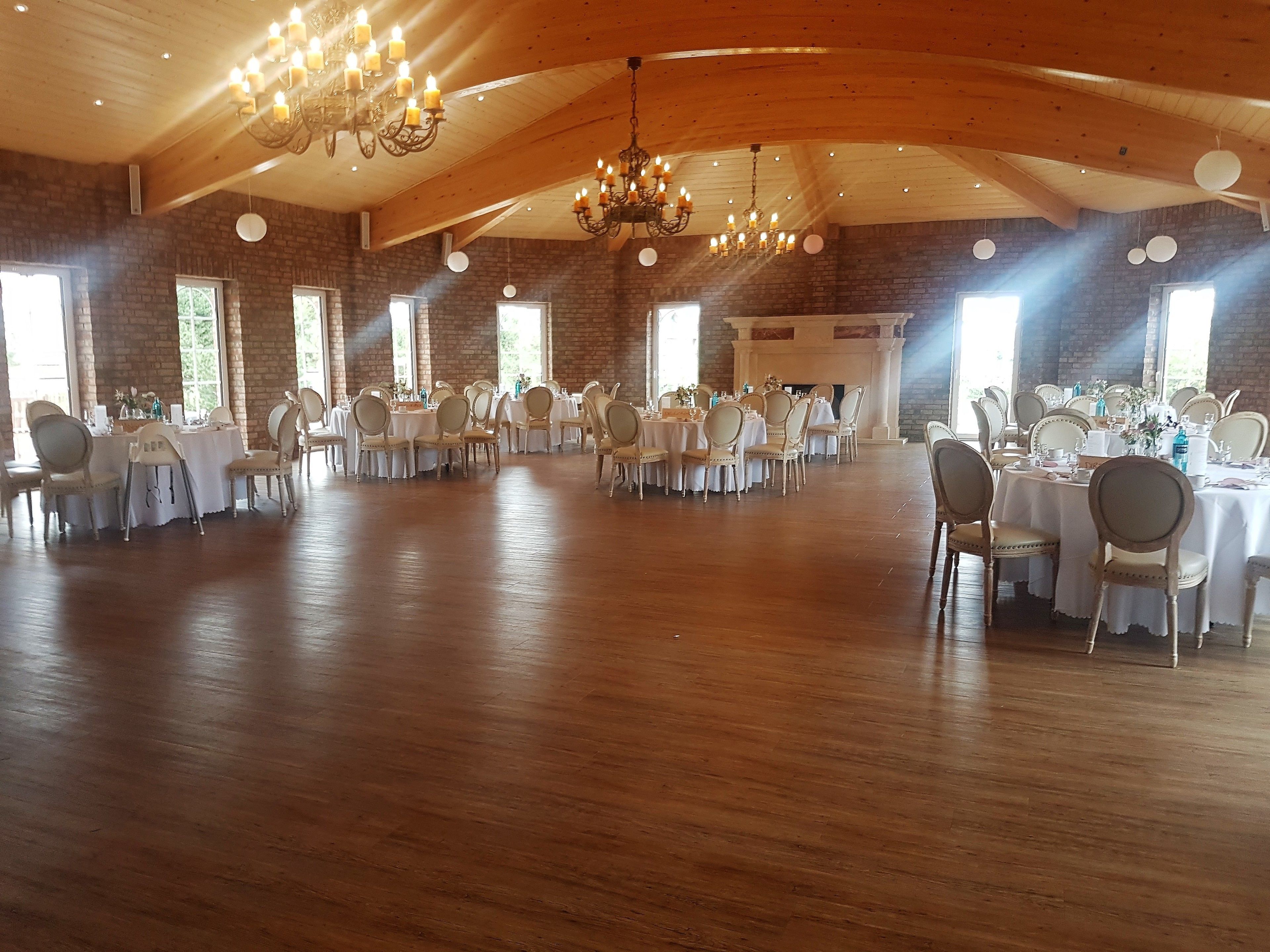 A large hall with multiple round tables and chairs, wooden floors, brick walls, and a high ceiling with chandeliers.