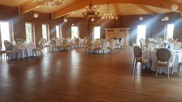 A large hall with multiple round tables and chairs, wooden floors, brick walls, and a high ceiling with chandeliers.