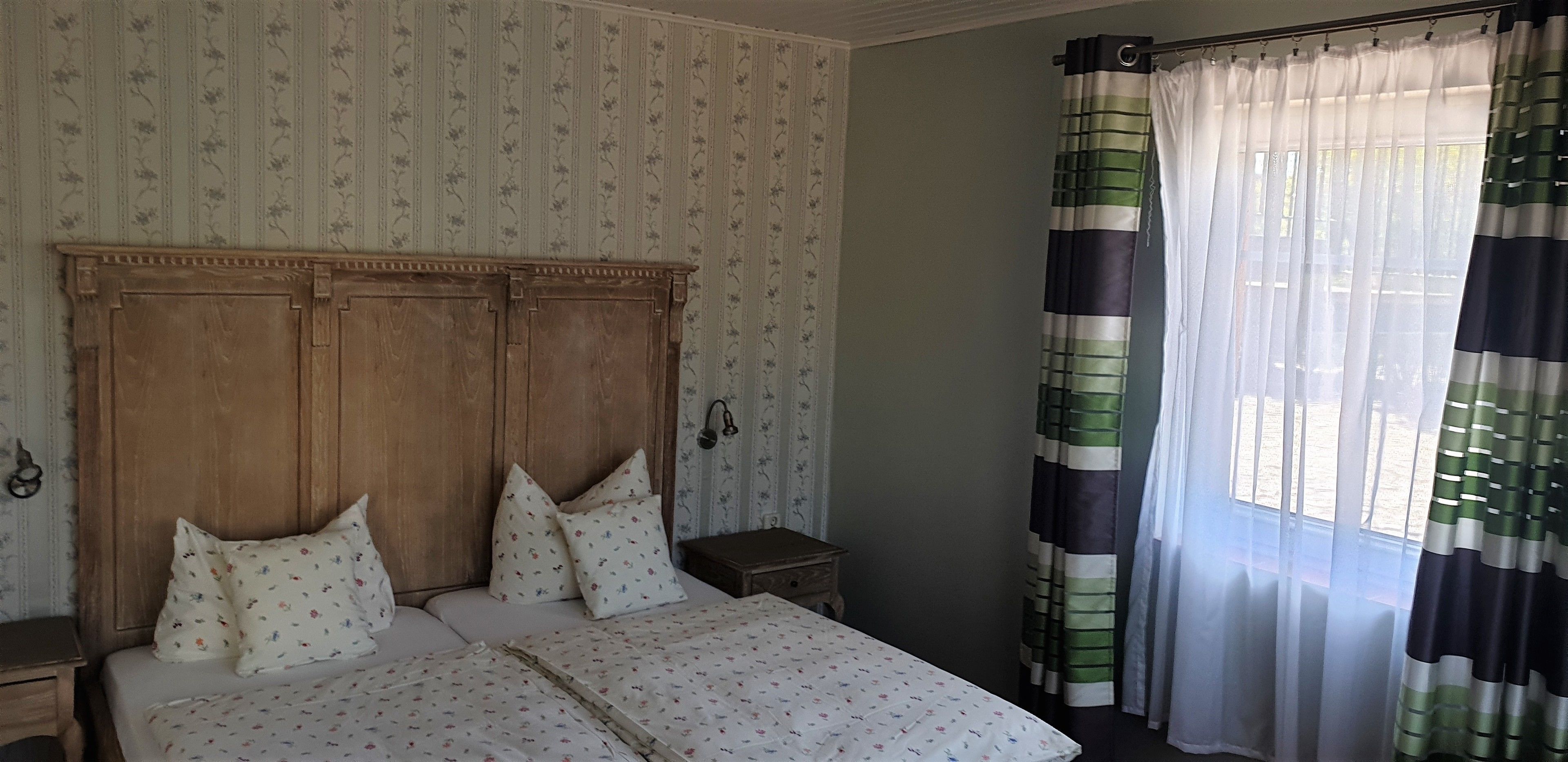 Camera da letto con testiera in legno, biancheria da letto a motivi floreali, un comodino e una finestra con tende a righe.