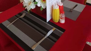 A dining area with a table set for two, a menu, condiments, and a vase with white flowers. - Nyaholoti Airport Hotel (Dar es Salaam)
