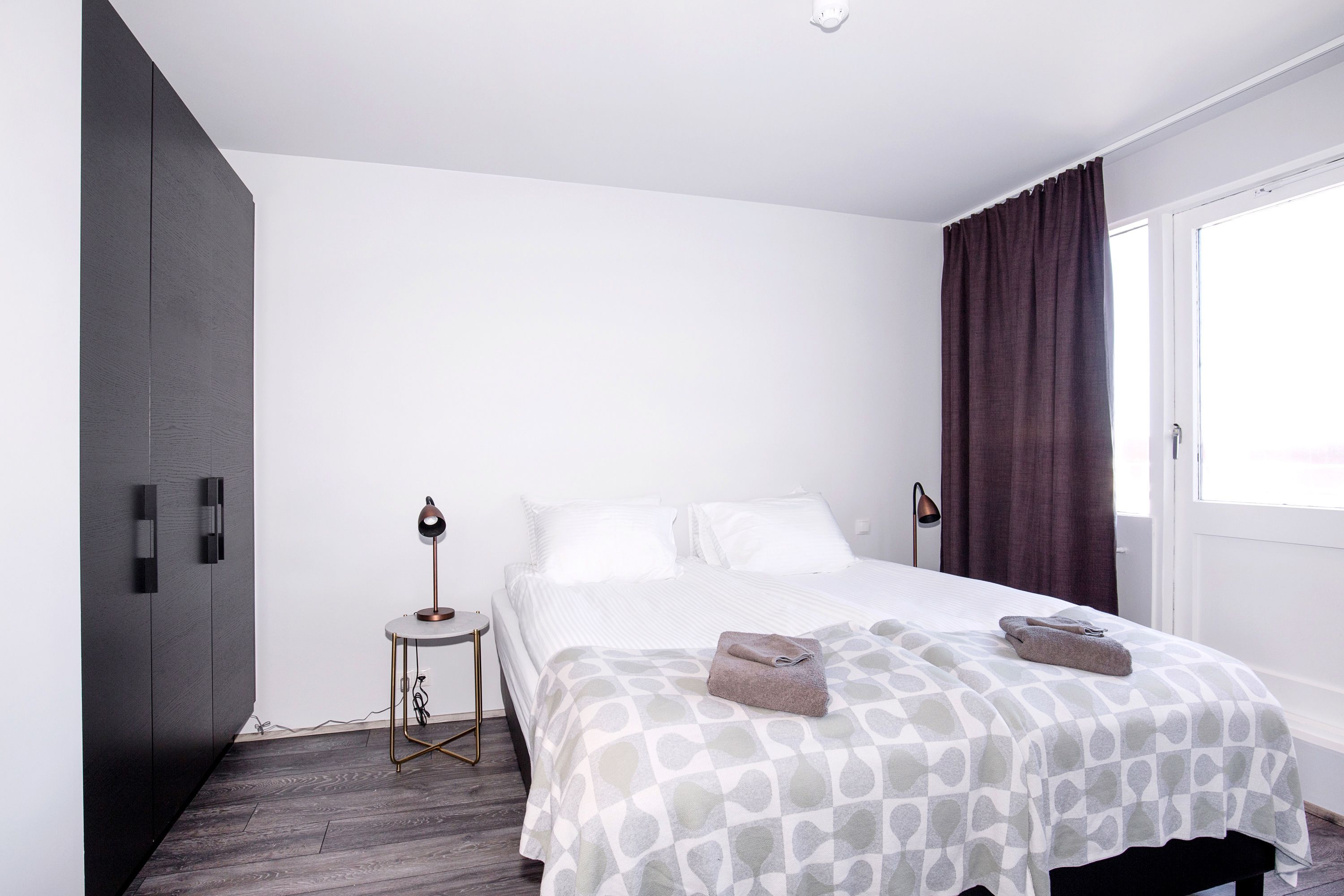 Een moderne slaapkamer met een groot bed, een donkere garderobe, nachtkastje met lamp en een raam met gordijnen.