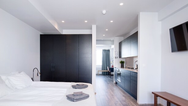 Quarto de hotel moderno com cama grande, armário escuro, pequena cozinha e um banco.