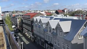 Vista da rua com edifícios, céu claro e uma torre ao longe.