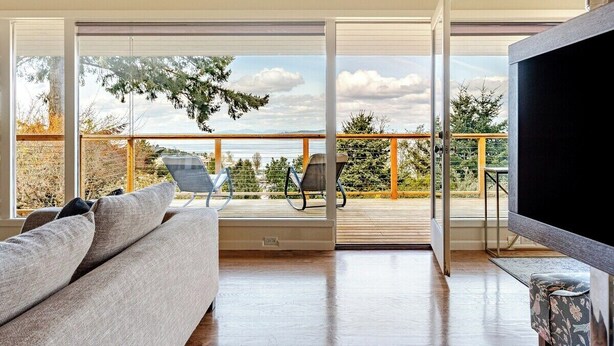 A modern living room with a sofa, a flat-screen TV, and a balcony with a view of the sky and trees. - Pritchard's Perch - private 2.5 acre in-city property, views from every room! (Seattle)