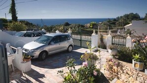 Zona residencial con un coche blanco estacionado en una entrada, rodeada de plantas en macetas y vista al mar.