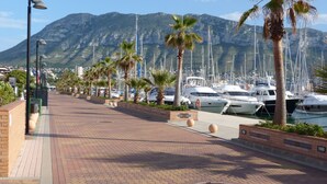 Ein Yachthafen mit vertäuten Booten, Palmen säumen die Promenade, und im Hintergrund Berge.